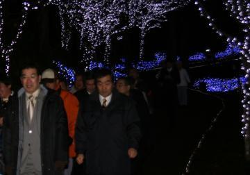 夜、暗い中、青や白い光で彩られたイルミネーションの中を知事と町長、関係者が歩いている写真