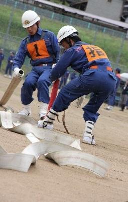 ヘルメットをかぶり活動服を着て胸にゼッケンを付けた消防団員の男性2名がホースを使った技術を披露している写真