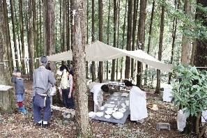 林の中の木に大きな白い布を貼って屋根を作っており、その下にシートを広げて作品が並べられており、来場者が作品を見ている様子の写真