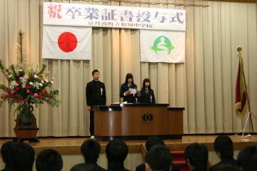 舞台上に「祝 卒業証書授与式」と書かれた横断幕と日の丸の国旗、京丹波町章が掲げられ、きれいに飾られた檀上花の横にある演台に3名の卒業生が立って、中央の女性が用紙を持って答辞を述べている写真