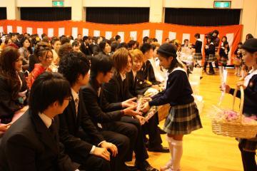 スーツ姿の新成人が会場内の席に座っており、子どもたちが、一人一人に一輪の花を手渡している写真