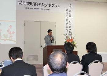 前方に「京丹波町観光シンポジウム」と書かれた横断幕があり、講演台に立つ井門隆夫さんがマイクを持って話をしている写真