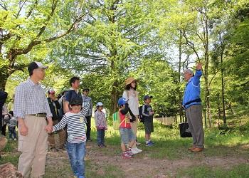 木々の生えた森の中で、紺色のジャンパーを着た男性が親子連れの参加者に説明をしている写真