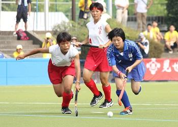白いユニフォームの選手と、青色のユニフォームの選手がスティックを持ち、白いボールを奪おうと競って走っている試合の様子の写真