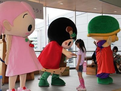 さくらちゃん、味夢くん、かめまるのマスコットキャラクターが立っており、味夢くんが髪の長い女の子と握手をしている写真