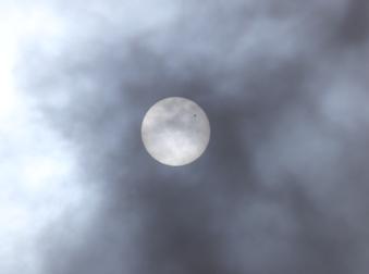 薄雲のかかった丸い太陽に小さく黒丸の影が太陽の右上に重なって見える写真