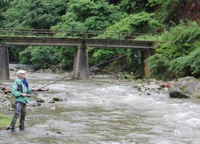 とおくに橋が見える山間にある川で、緑色のジャンパーを着た男性が川岸に立って、川に竿を下ろして釣りをしている写真