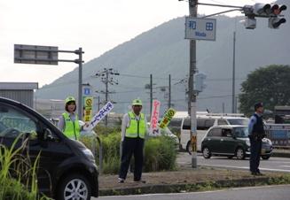 黄緑色の帽子とベストを着た役員の男性と女性が交差点前の歩道に立ち、街頭啓発をしている写真