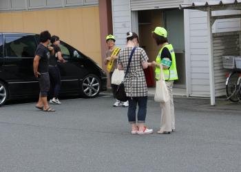 黄緑色の帽子とベストを着た役員の女性が、町民の女性に啓発物品を手渡し、話をしている写真