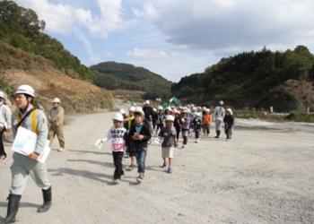 周りがが小高い丘のようになっており、乾燥したダム湖の底を作業服を着た職員の後をついて歩いている下山小学校の児童達の写真