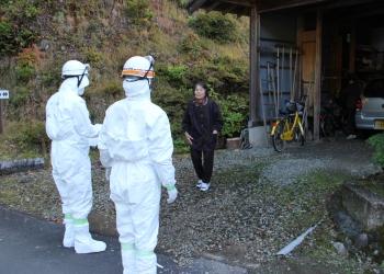 白いヘルメットを頭に被り、全身白い防護服を着た2名の消防団員の方が、住民の年配の女性避難を呼び掛けている訓練の様子の写真