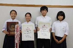 優勝盾、トロフィー、賞状を持っている3名の女子生徒と1名の男子生徒が笑顔で並んで写っている写真