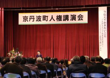 前方舞台上に「京丹波町人権講演会」と書かれた横断幕、シナリオライターの丘乃れいさんが舞台前で話をしており、会場内には沢山の来場者が席に着いて、講演を聞いている様子を後方から写した写真