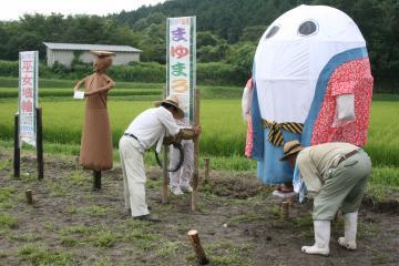 まゆまろや巫女埴輪(みこはにわ)の看板の横に、手作りの案山子を設置する作業を男性2名が行っている写真