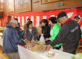 紅白の幕が壁に掛けられた会場内で、台の上に農産物が陳列され、訪れた来場者が台の周りに集まって話をしている写真