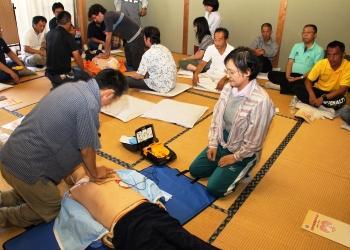 訓練用の人体モデルの胸に、AEDの電極パッドを貼り、男性がヒジを伸ばし手を重ねて人体モデルの胸に置き、胸骨圧迫をしており、その様子を参加者の方々が座って見ている講習会の様子の写真