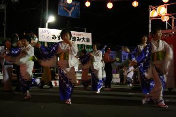 全体が白で右肩と裾が青紫色の柄の着物を着た踊り子の皆さんが左脚を挙げて両手を顔の高さまで挙げて踊りを踊っている写真