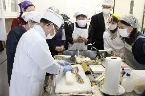 白いコックシャツを着た男性がまな板の上で調理しており、周りに参加者が集まって料理の手順を見ている写真