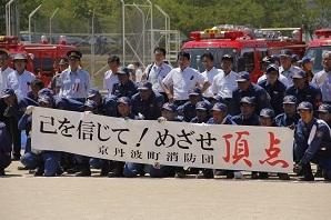 「己を信じて!めざせ頂点 京丹波消防団」と書かれた横断幕を持って、消防団の方々が、消防車両の前に集まって写っている集合写真