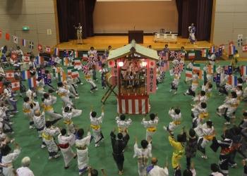 会場内の中央に提灯がともり、紅白幕が巻かれた櫓が組まれ、櫓を囲んで円を作り、浴衣を着た踊り子の皆さんが、両手を上にあげて踊りを踊っている写真