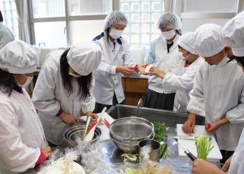 白い帽子、マスク、割烹着を着た生徒たちが調理台のまな板の上で、食材を切ったり、友達の作業の様子を見守ったりしている調理実習の様子の写真