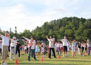 陸上競技場に、等間隔に広がった大勢の参加者が、両手を斜め上に挙げてラジオ体操をしている写真