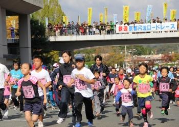 胸にピンク色のゼッケンを付けた子どもや大人、女性、男性など沢山のランナーが走っており、後方の橋の上には、のぼり旗や横断幕がかけられ、沢山の観客が応援に来ている「2012京都丹波ロードレース」の様子の写真