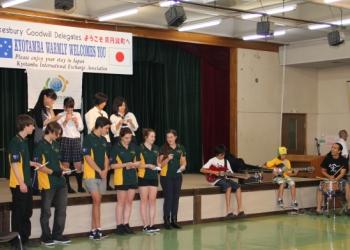 緑色のポロシャツを着た留学生と生徒の皆さんが2列に並んで立ち、2名の生徒がギターを演奏して歌を披露している写真