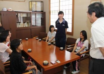 6名の派遣生がテーブルの席に着き、中央に座る女性が席から立って話をしており、町長が笑顔で話を聞いている写真