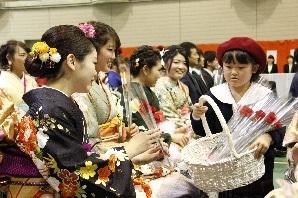 赤いベレー帽を被った女の子が赤い一輪ブーケの花が沢山入ったかごを持って、艶やかな振袖を着た新成人の方々に花を渡している写真