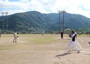 遠くに山が見える広々とした球場で、バッターボックスに立ち、バットを振り切った打者と、グラウンドに広がって守備をしている選手たちが写っているソフトボール大会の様子の写真
