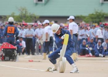 沢山の消防団員の方々が遠くで見学をしており、ヘルメットをかぶり、活動服を着た消防団員が消防用のホースを片足で抑え、両手でホースを延長する作業を方法を披露している写真
