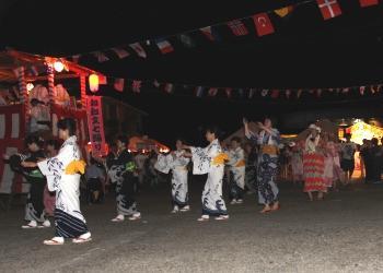 提灯が灯され、紅白幕が巻かれた櫓、万国旗が飾られた薄暗い夜の会場、浴衣を着た沢山の参加者が踊りを踊っている写真