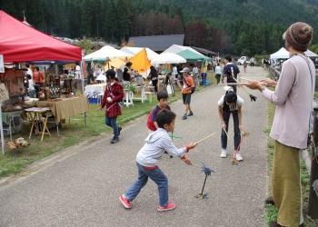 小道で子ども達が短い棒を使ったおもちゃで遊んでおり、公園内に沢山の色とりどりのテントが立ち並んでいるフリーマーケットでにぎわう会場の様子の写真