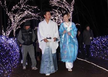 白い紋付きと青のグラデーションの袴、鮮やかな水色の花嫁衣装を着た夫婦が、青と白色の光のイルミネーションで彩られた夜の道を笑顔で歩いている写真
