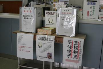 机の上に置かれた東日本大震災義援金用の募金箱と、支援物資受付場所を示す矢印が記載された張り紙が貼ってある写真