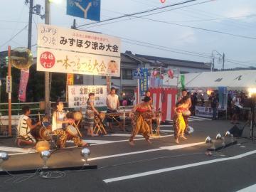 みずほ夕涼み大会の看板の前で、照明でライトアップされながら太鼓の演奏を行っている出演者の写真