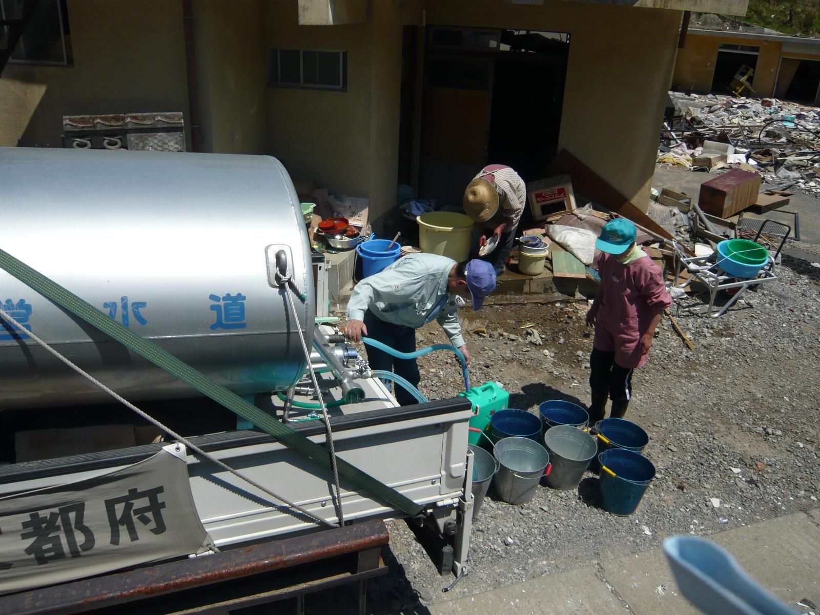 家財を洗う市民のため、半壊家屋に給水車両で給水を行う職員の写真