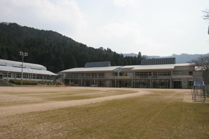 広い運動場の脇にうんていやジャングルジムなどの遊具がいくつか設置され、奥にはL字型に校舎が並んでいる和知小学校の外観写真