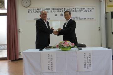 調印式で片手で握手し、もう片方の手で調印した協定書を一緒に持っている松本教育長と畠中副理事長の写真