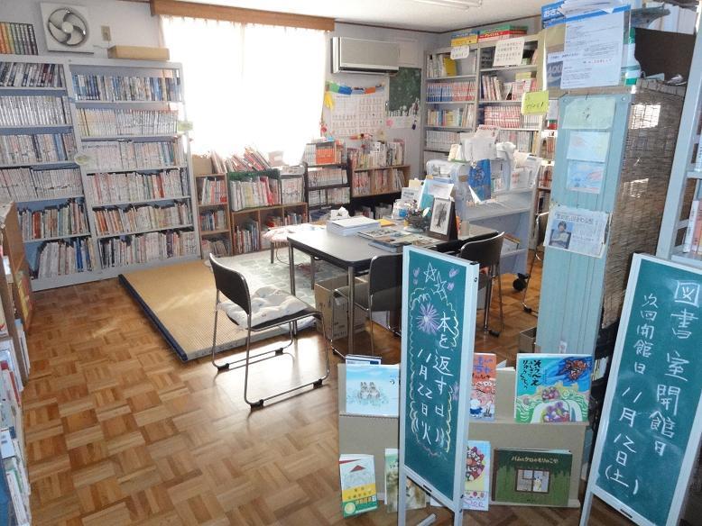 室内の壁側に沢山の本が立てられた本棚が並んでおり、畳の敷かれたスペースと机と椅子設置された図書室内の写真