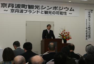 「京丹波町観光シンポジウム 京丹波ブランドと観光の可能性」と書かれた横断幕の前の演台で寺尾町長が話をしており、沢山の方々が座って話を聞いている講演会の様子の写真