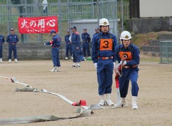 ヘルメットを被り、紺色の活動服、胸にゼッケンを付けた消防団員の1名が直立して立ち、その横で1名が消防のホースを両手で持ち、左脚を引いて腰を少し落とした姿勢で構えている訓練の様子の写真