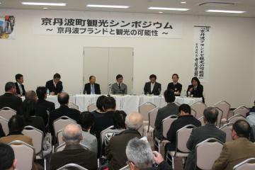 前方の長机に関係者の方々8名が座り、意見を述べ討論しており、沢山の参加者の方々が椅子に座って話を聞いているパネルディスカッションの写真