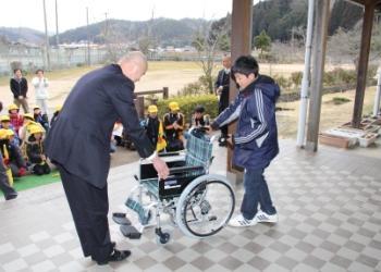 真新しい紺色と緑色のチェック柄の車椅子を代表の男子児童が紺色のスーツを着た男性に贈呈しており、その様子を他の児童が見守っている写真