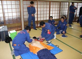 紺色の活動服を着た隊員が訓練用の人体モデルを使って、気道確保、AEDの機械の装置を操作をしている。その様子を後方から見ている隊員の写真