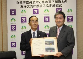 「京都府京丹波町と北海道下川町との友好交流に関する協定締結式」と書かれ、京丹波町町章と下川町章が交互にデザインされたボードの前で、寺尾町長と北海道下川町の安齋町長が協定書を持って並んで写っている写真