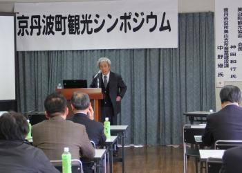 「京丹波町観光シンポジウム」と書かれた横断幕の前で、講演台の前に立って話をしている神田会長と長机の席に着いて話を聞いている参加者の方々を後方から写した写真