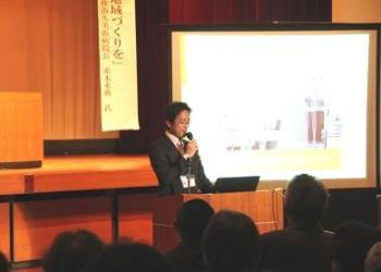 前方の大きなスクリーンに資料が映され、講演台に立っている一人のスーツを着た男性がマイクを持って話をしている写真