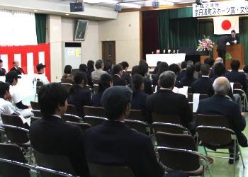 前方の舞台に横断幕と日の丸が掲げられており、講演台の前に立って賞状を受け取り表彰を受けている一人の男性、その様子を見守っている参列者の方々を後方から写した写真